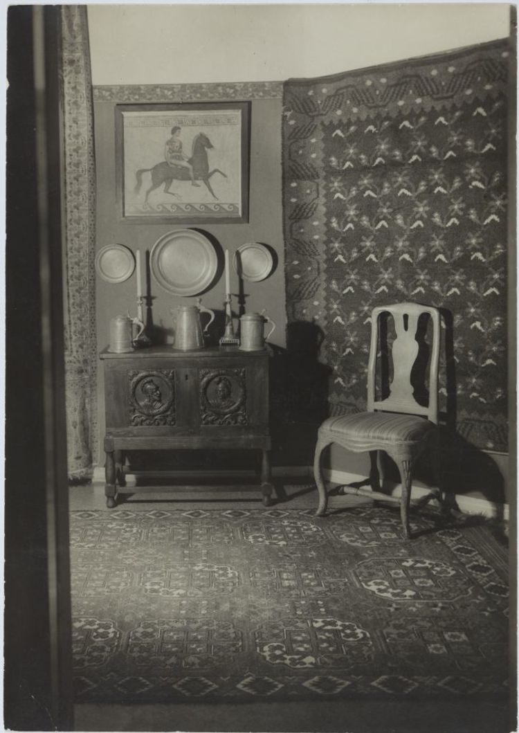 A chair beside a engraved sideboard. On top of the sideboard three pitchers and two candle sticks. Three plates on the wall.
