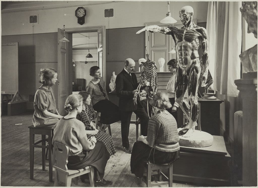 Man teaching six students anatomy in class, with a life sized human sculpture and skeleton as example.