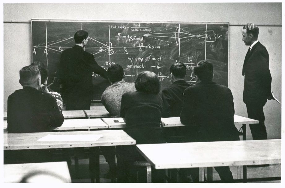 Two men standing in a class room drawing on a chalk board with six students sitting and watching.