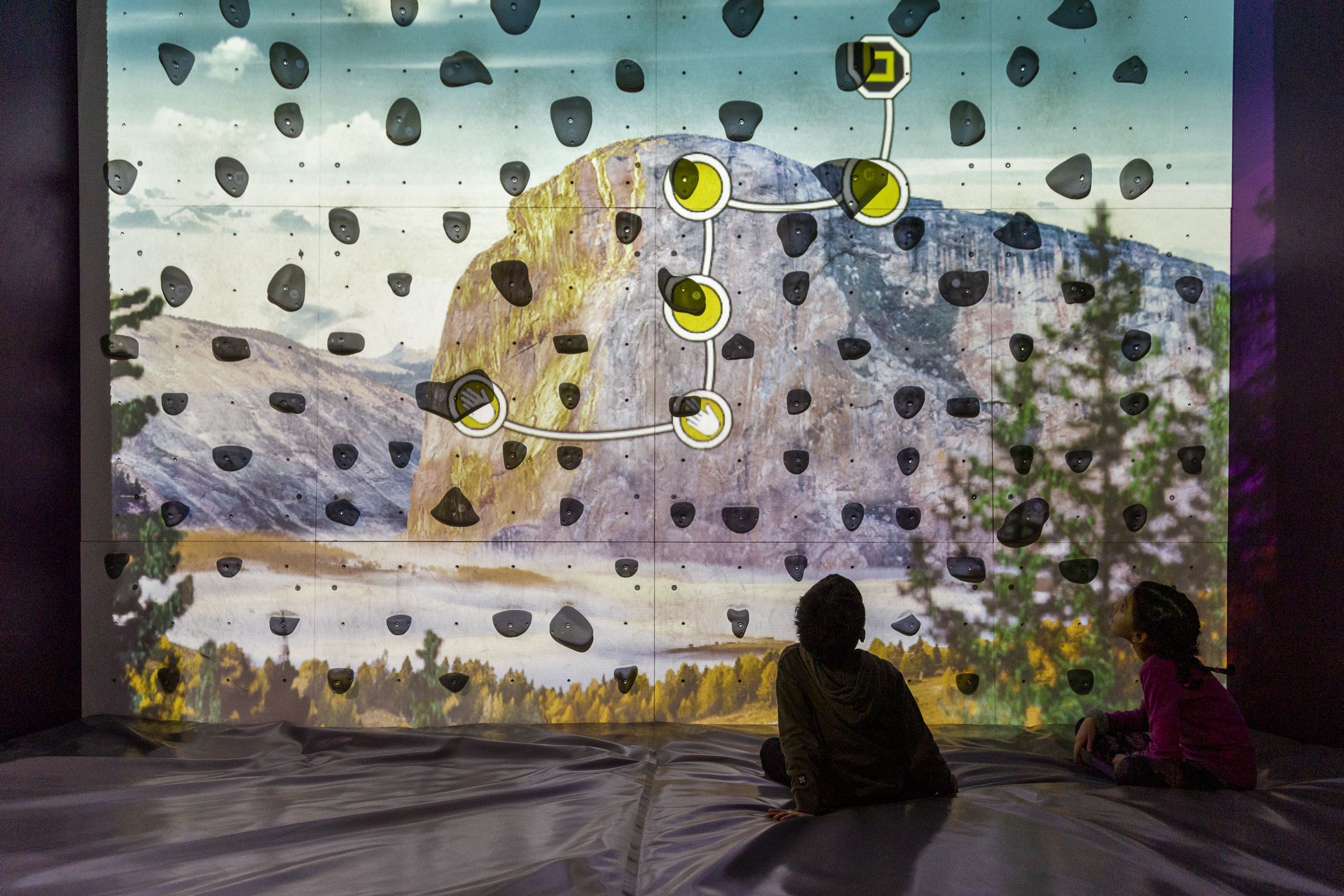 A photo of a climbing wall with a projection on it. The young boy and girl are sitting at the bottom right of the photo, looking up at the projection. Their posture suggests that they are tired. The projection shows a granite mountain in the background, perhaps from Yosemite, some mist near the ground in the middle distance, and trees in the foreground, out of focus on the left and right, framing the scene. A climbing route is also projected onto the wall: two green circles with hand icons are the start of the route, around the center of the photo; then white lines connect the right hand in sequence to two green circles above it; then the route turns right and continues out of the photo. Each circle is projected onto a climbing hold on the wall.