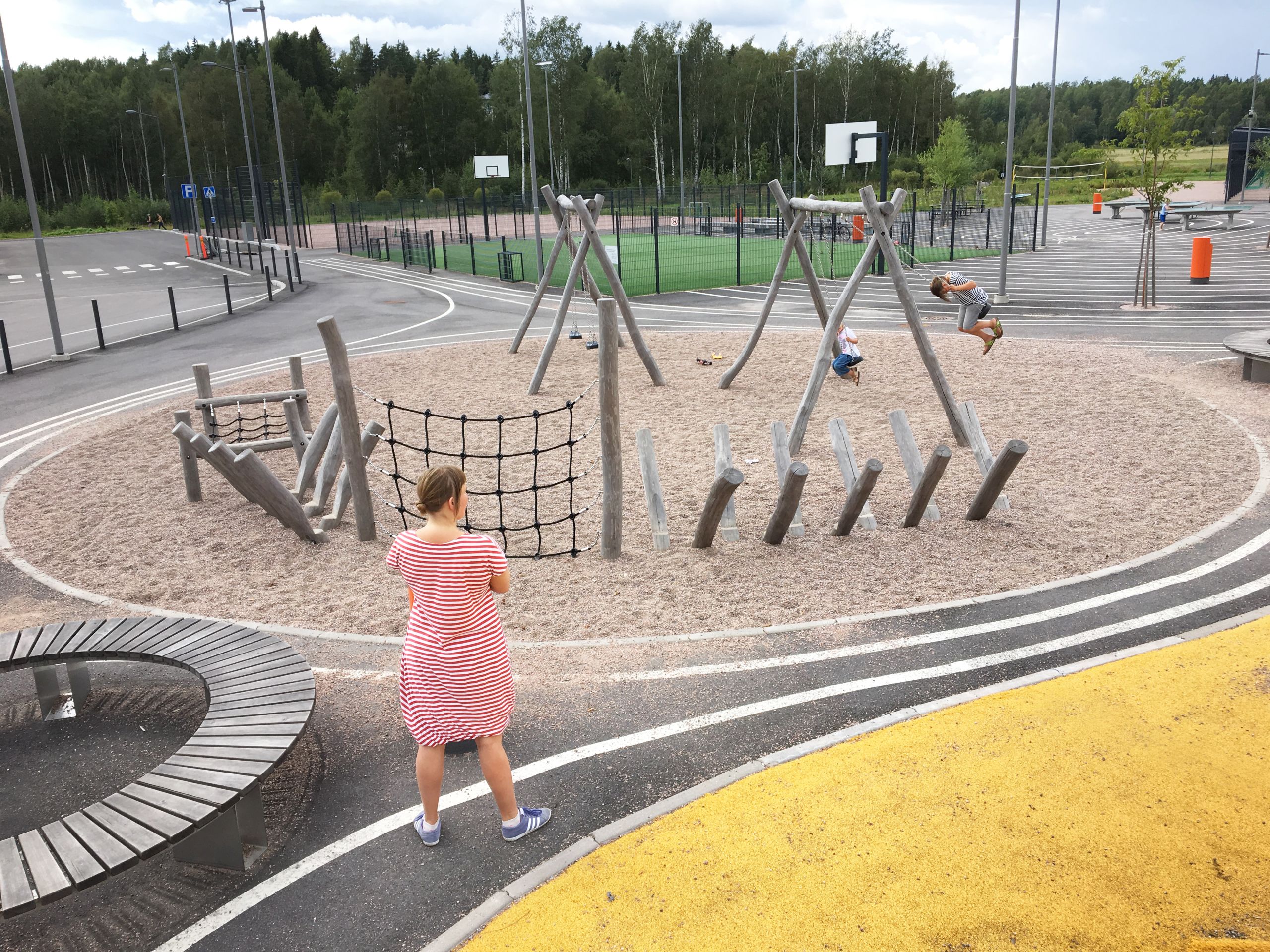 A parent supervising a child in a swing on a playground with wooden structures and sand. 