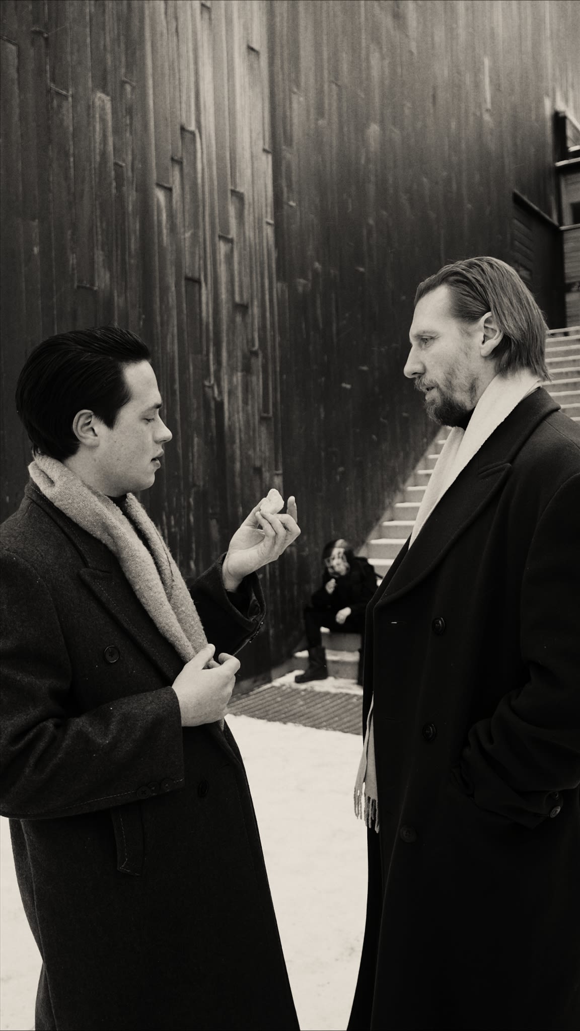 two men talking to each other with a person sitting on the stairs in the background